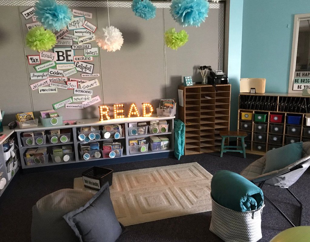 Cozy classroom reading nook with bean bag chairs and cushions, fostering a student centered learning climate. Shelves brim with bins of books labeled by genre. READ is spelled out in lit letters, while motivational words embellish the walls. Soft lighting and colorful pom-poms enhance the welcoming atmosphere.