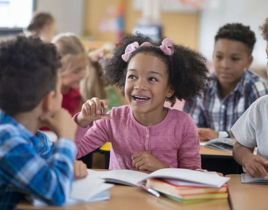 A young girl with curly hair and a pink sweater smiles and talks with classmates, practicing classroom management strategies. She is surrounded by other children, open books on their desks. The classroom buzzes with brightness and activity.