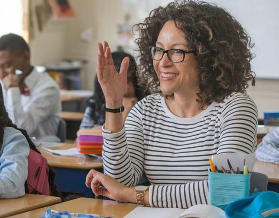 A woman with curly hair and glasses smiles as she raises her hand in a classroom filled with students and supplies. Sporting a striped shirt, she exudes confidence, embodying effective classroom management strategies while seated at her desk.