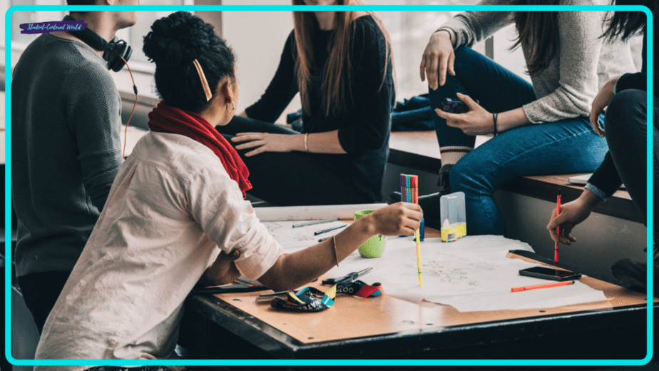 A small group of people is sitting and collaborating around a table with papers, pens, and a smartphone. One person has headphones around their neck, and another holds a pencil near the paper. They appear to be working on a project or study task.