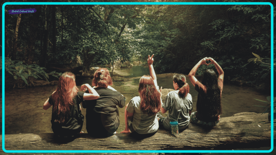 A small group of five people sits on a log by a forest stream, facing away while making hand gestures like peace signs and a heart. The scene is surrounded by lush greenery and dappled sunlight.