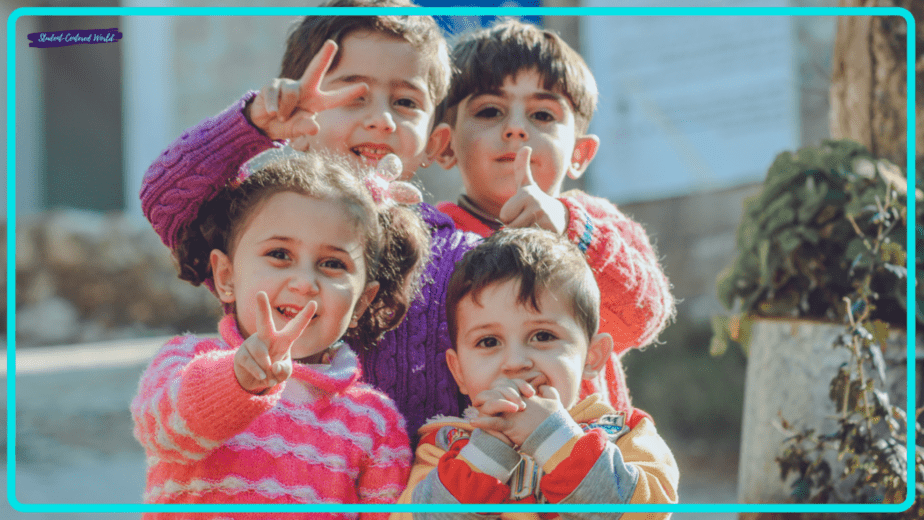 A small group of four children smile and make peace signs towards the camera. Dressed in colorful sweaters, they appear to be enjoying a sunny day outdoors, with trees and a building visible in the background.