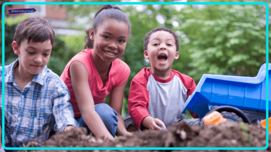 In a small group, three children play happily in a garden. A girl in a red shirt kneels between two boys. One boy is wearing a plaid shirt, and the other in a red and gray shirt smiles, holding a toy truck. Greenery surrounds them.