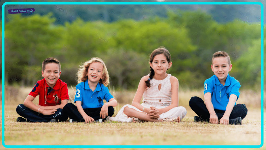 A small group of four children sits on the grass in a park, beaming at the camera. Dressed in bright colors, two boys wear red and blue shirts, another boy is also in blue, while a girl wears a white dress. Lush green trees and rolling hills provide a picturesque backdrop.