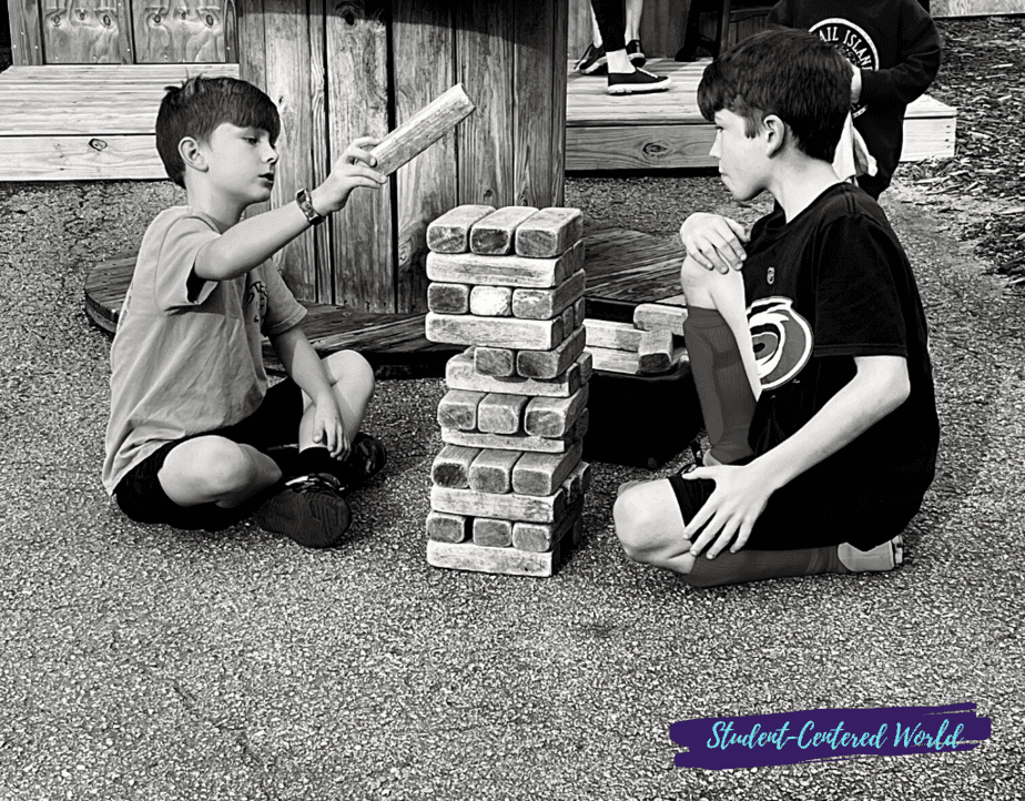 Two boys, part of Generation Alpha, sit on the ground, engrossed in playing a giant Jenga game with large wooden blocks. One reaches to place a block on top. A Student-Centered World graphic graces the bottom right corner.