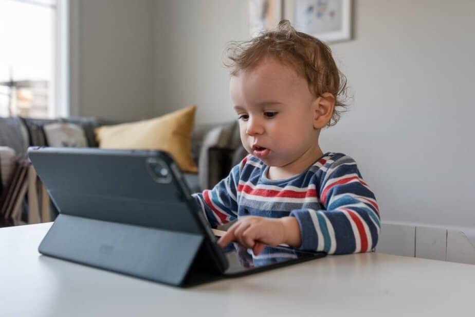 A toddler with curly hair is sitting at a white table, focused on using a tablet in a gray case. The child is wearing a striped shirt, and theres a sofa with cushions in the background.