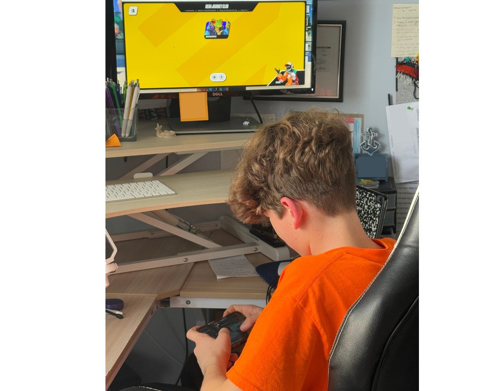 A person in an orange shirt is sitting at a desk and playing a game on a computer monitor. They are holding a controller, focused on the yellow game screen. The desk has papers, a keyboard, and a notepad.